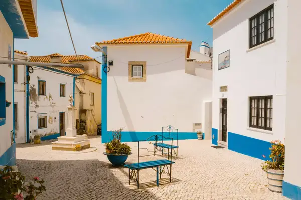 A sunny courtyard surrounded by white buildings with blue accents, featuring benches and potted plants