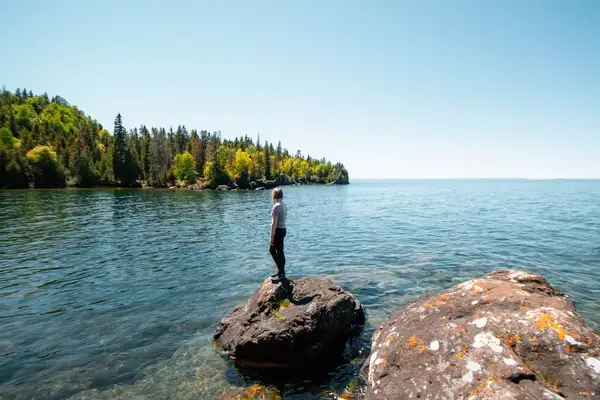 A woman stands on a rock with her back to the camera, looking over the water to trees in the distance