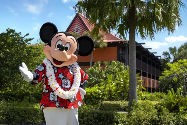 mickey mouse mascot wearing Lei standing with hand out in front of outdoor scene of palm tree