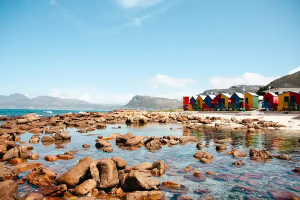 Coastal landscape featuring colorful beach huts in Muizenberg, Cape Town, with rocky shoreline and mountains in the background