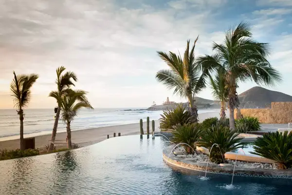 Beachfront scene with palm trees, an infinity pool, and an ocean view, with distant sand dunes and structures on the horizon