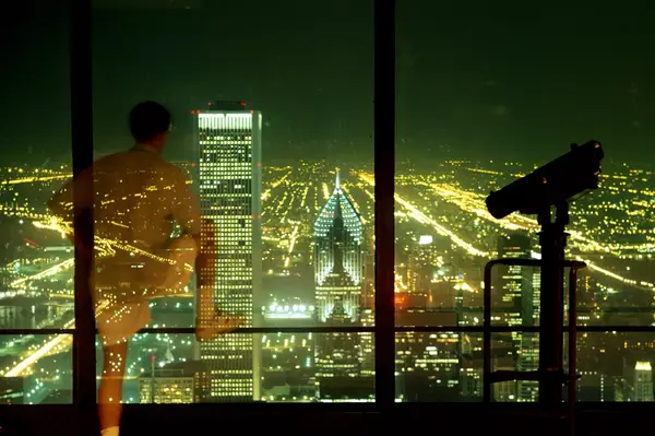 A man looks out through an observation deck window over the skyline of Chicago at night