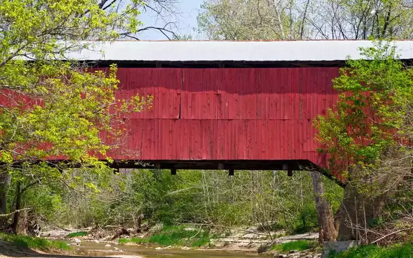 One of the many red-sided covered bridges of Parke County, Indiana, is seen standing over a tree-lined creek.