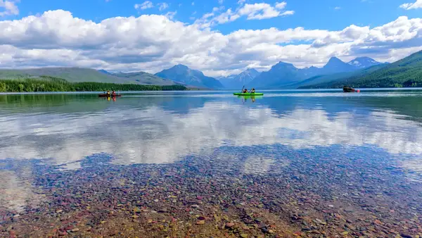 Kayakers on clear lake with colorful pebbles and mountains in the background