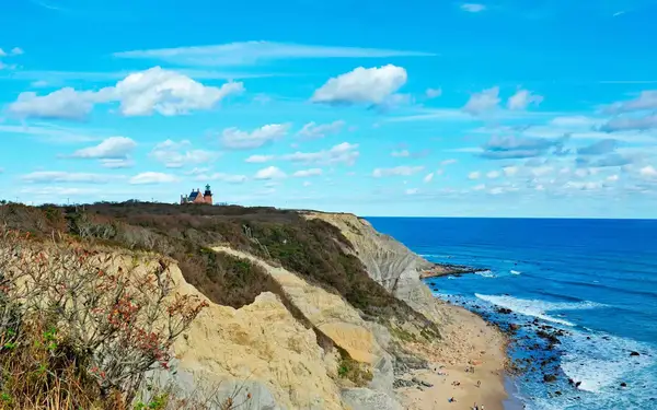 The rocky, moss-covered Mohegan Bluffs stand at the edge of a stretch of blue ocean water in Rhode Island, with a large lighthouse and home silhouetted in the distance.