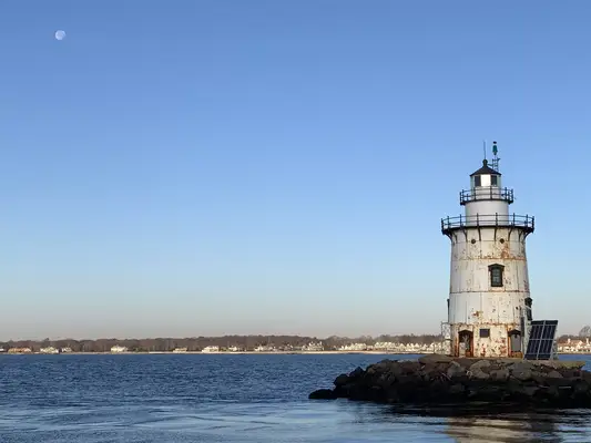 The Saybrook Breakwater Lighthouse stands in the foreground of a waterfront photo with the early-morning moon illuminated in the sky