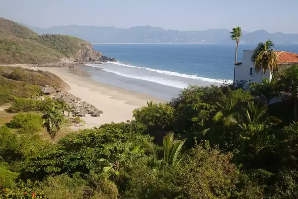 Jungle in foreground; red tile roof building, waves, ocean and mountains beyond
