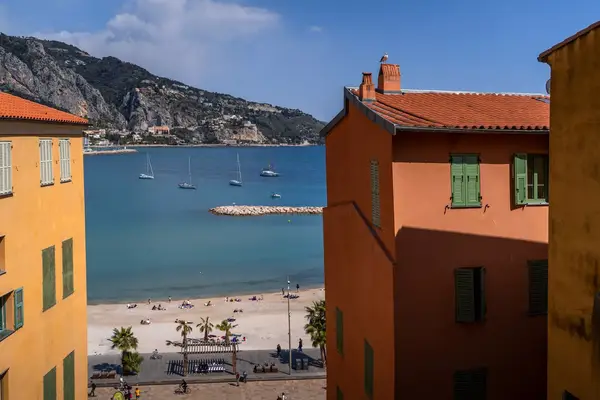 A coastal town view framed by buildings, with a beach, palm trees, and sailboats in the distance