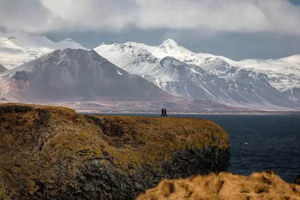 Two people stand on a cliff with a scenic backdrop of mountains