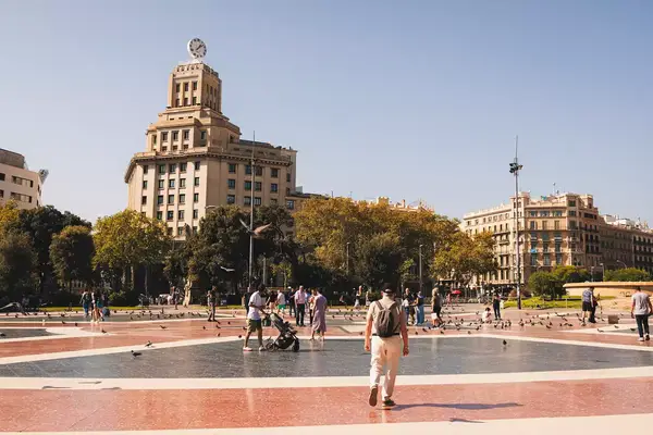 People walking in an urban plaza with a large historic building and trees in the background