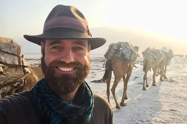 A person smiling in a desert setting with camels carrying cargo in the background