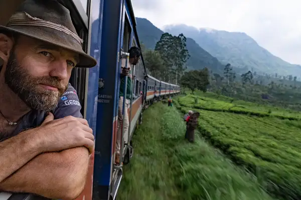 A person leaning out of a moving train window, scenic landscape with mountains and greenery in the background