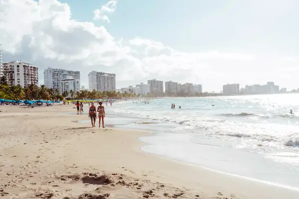 Two women walking down the beach in San Juan with sky scrapers behind them