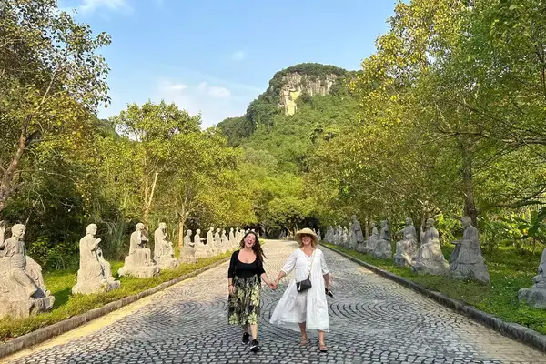 Mother and daughter hold hands walking down a pathway lined with statues and trees