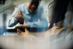 Close up of a salesman giving new car keys to his black customers in a car showroom. The view is through glass.