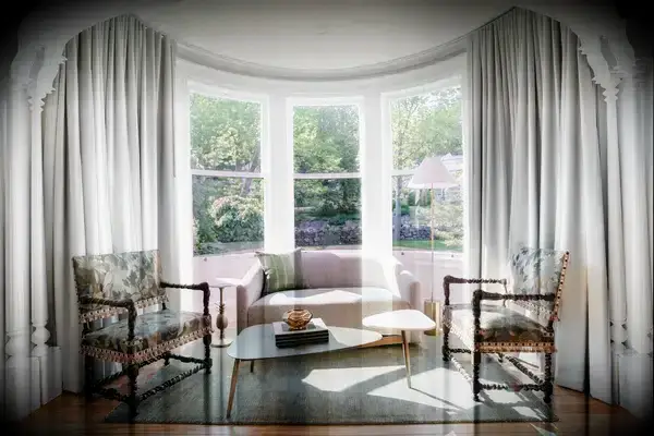 A couch, coffee table, and two armchairs in a room with large bay windows overlooking a green outdoor scene