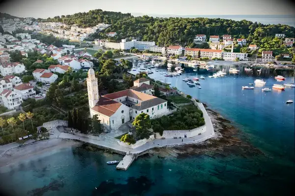 A coastal town featuring a historic church on a peninsula with housing and greenery in the background, surrounded by calm waters with boats