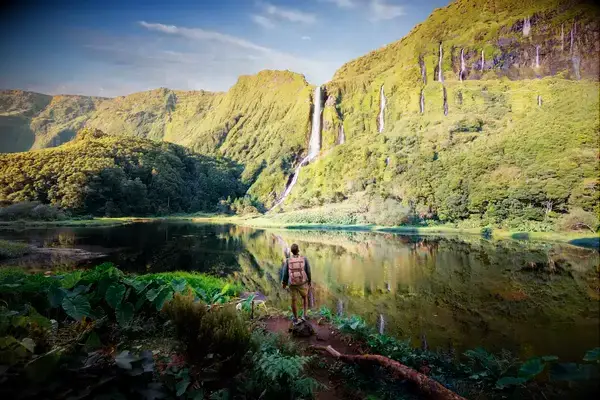 Person with a backpack standing in front of a lake and a distant waterfall among green hills