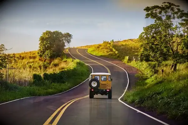 A vehicle driving along a hilly, winding road surrounded by greenery and trees, with a clear sky above