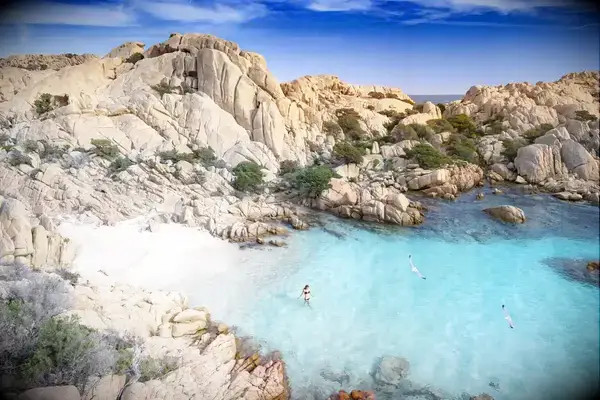 A scenic coastline with rocky cliffs and turquoise waters, a person wading and paddleboarders visible in the water