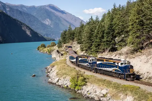 A scenic train traveling alongside a lake and tree-covered hills with mountains in the background