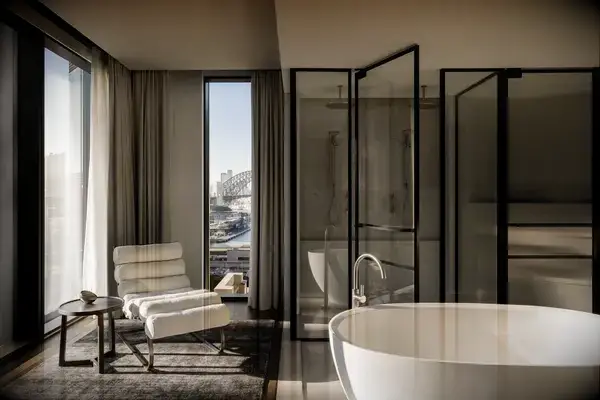 A modern bathroom suite with a freestanding bathtub, a chair by the window, and a view of Sydney Harbor Bridge in the background