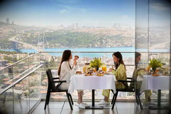 Two women dining at a table on a terrace with a view of a bridge and cityscape in the background