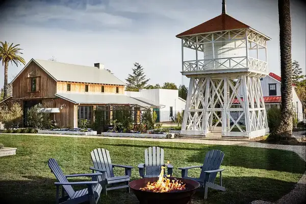 Outdoor scene with wooden barn, white tower structure, chairs around a fire pit, and palm trees on grass