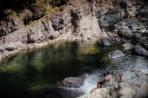 A man sits on rocks next to a natural pool surrounded by rocky terrain, with another person visible in the water