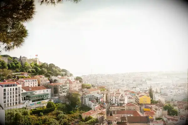 Overview of Lisbon, Portugal, cityscape featuring buildings and a bridge in the background, greenery visible in the foreground, sunny day