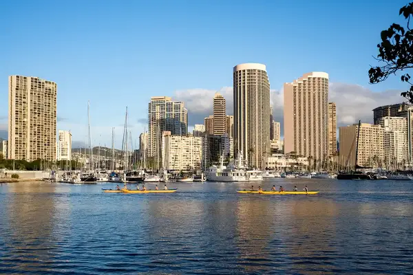 Skyline of a city with tall buildings and a marina, viewed across a body of water with small boats visible